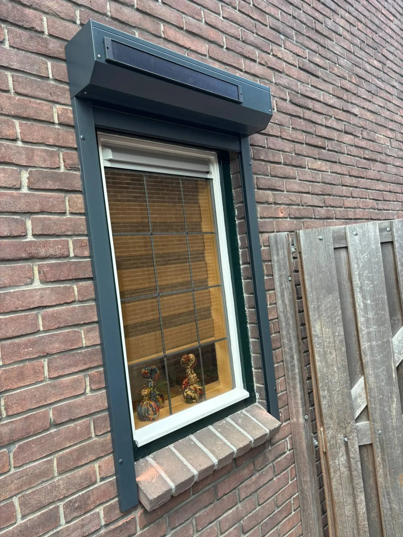Rolluikkast met een smal zonnepaneel erop, boven een raam in een bakstenen gevel